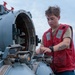 A person wearing a red vest and tan shirt is working on what appears to be a naval weapon system or torpedo tube aboard a military vessel at sea. The individual has short brown hair, visible tattoos on their forearm, and is wearing a watch while manipulating components of the equipment. The ocean and overcast sky are visible in the background.