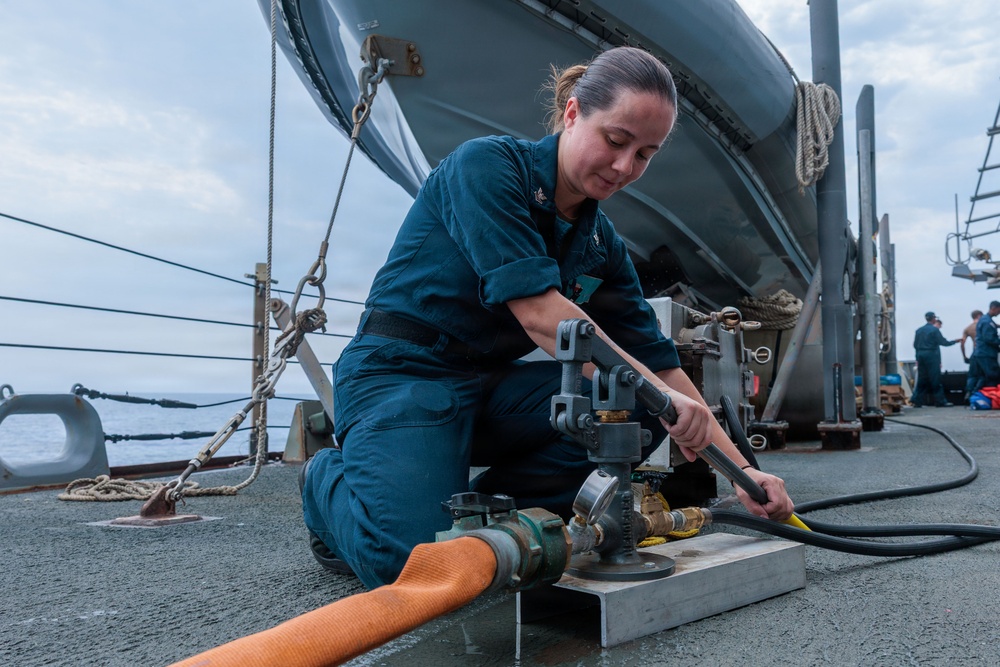 A female sailor in dark blue Navy coveralls is kneeling on the deck of a naval vessel, working with hose connections and valve fittings. She appears to be conducting damage control or firefighting equipment maintenance, connecting or adjusting hoses at a deck fitting. The scene is set on an open ship deck at sea, with a lifeboat and other crew members visible in the background.