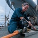 A female sailor in dark blue Navy coveralls is kneeling on the deck of a naval vessel, working with hose connections and valve fittings. She appears to be conducting damage control or firefighting equipment maintenance, connecting or adjusting hoses at a deck fitting. The scene is set on an open ship deck at sea, with a lifeboat and other crew members visible in the background.