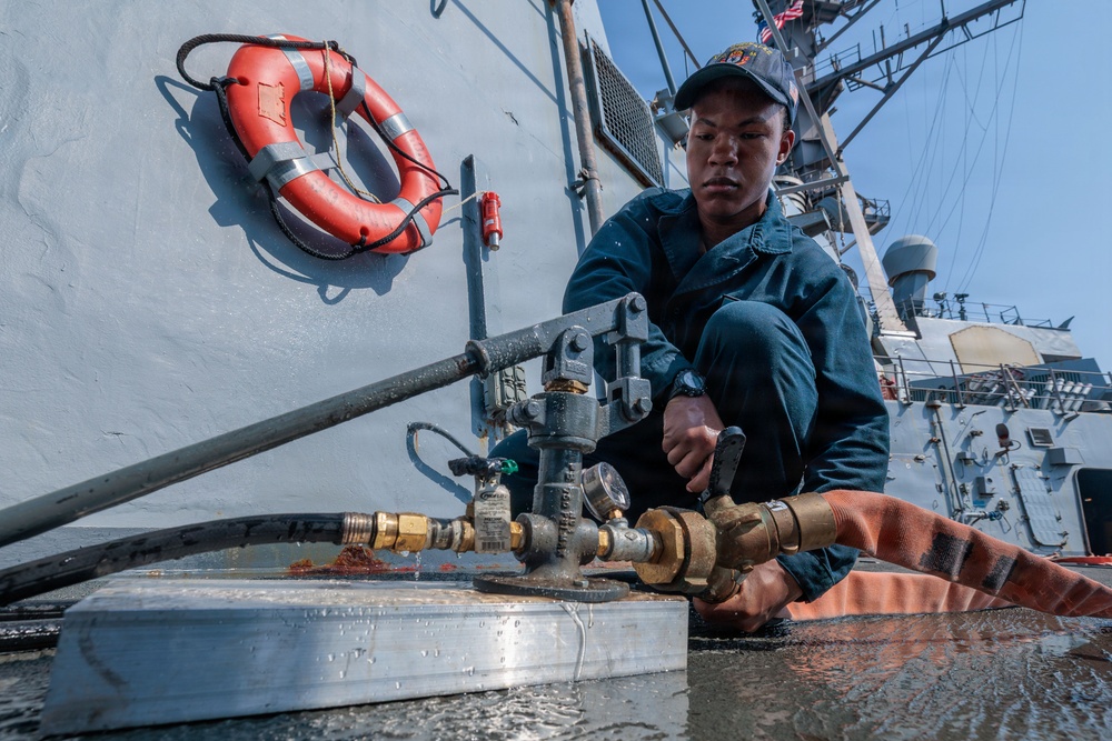 A sailor in dark blue coveralls and a cap is crouched on the deck of a naval vessel, working with hose connections and valve fittings. The low-angle photograph emphasizes the plumbing hardware in the foreground, with a life ring mounted on the ship's hull visible in the background. The setting appears to be the exterior deck of a U.S. Navy warship, likely a destroyer or cruiser.