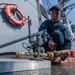 A sailor in dark blue coveralls and a cap is crouched on the deck of a naval vessel, working with hose connections and valve fittings. The low-angle photograph emphasizes the plumbing hardware in the foreground, with a life ring mounted on the ship's hull visible in the background. The setting appears to be the exterior deck of a U.S. Navy warship, likely a destroyer or cruiser.