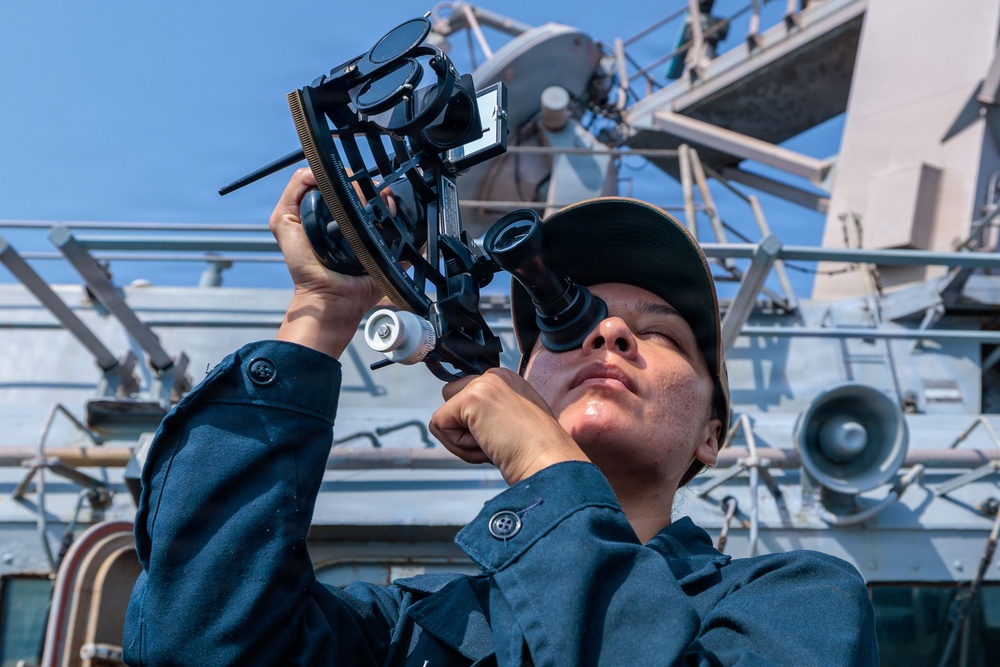 A naval personnel member in dark blue uniform and cap is using a sextant aboard a military vessel. The individual is looking through the eyepiece of the sextant, likely performing celestial navigation. The image is taken from a low angle on the ship's deck with naval equipment and superstructure visible in the background.