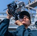 A naval personnel member in dark blue uniform and cap is using a sextant aboard a military vessel. The individual is looking through the eyepiece of the sextant, likely performing celestial navigation. The image is taken from a low angle on the ship's deck with naval equipment and superstructure visible in the background.