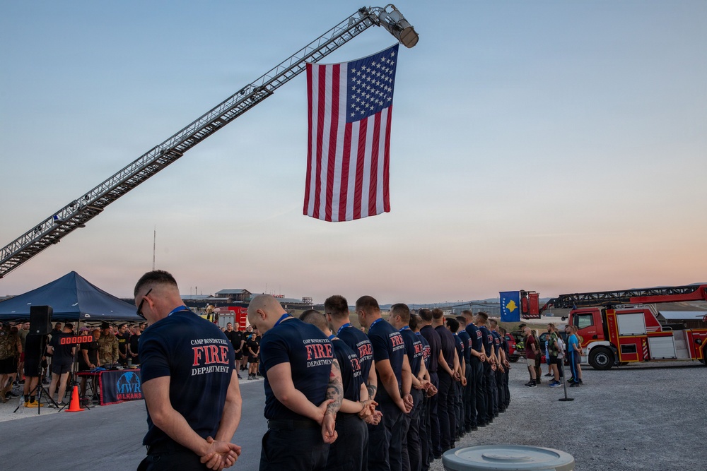 A group of firefighters from Camp Bondsteel Fire Department stand in formation with heads bowed during what appears to be a memorial or ceremonial event. A large American flag is suspended from an extended fire truck ladder above the assembled personnel. The event appears to take place at dusk in an outdoor area consistent with a military installation.