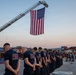 A group of firefighters from Camp Bondsteel Fire Department stand in formation with heads bowed during what appears to be a memorial or ceremonial event. A large American flag is suspended from an extended fire truck ladder above the assembled personnel. The event appears to take place at dusk in an outdoor area consistent with a military installation.
