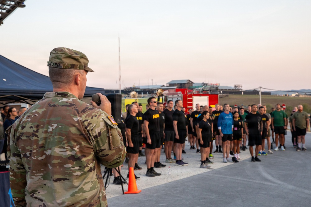 A U.S. Army soldier in OCP (Operational Camouflage Pattern) uniform, identified by the name tape 'LACOUNT', addresses a large formation of personnel wearing black Army PT (Physical Training) uniforms. The event appears to be an outdoor military physical fitness formation or ceremony, taking place on what looks like a tarmac or airfield area at dawn or dusk. A red fire truck is visible in the background, suggesting proximity to an airfield or installation.