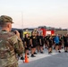 A U.S. Army soldier in OCP (Operational Camouflage Pattern) uniform, identified by the name tape 'LACOUNT', addresses a large formation of personnel wearing black Army PT (Physical Training) uniforms. The event appears to be an outdoor military physical fitness formation or ceremony, taking place on what looks like a tarmac or airfield area at dawn or dusk. A red fire truck is visible in the background, suggesting proximity to an airfield or installation.