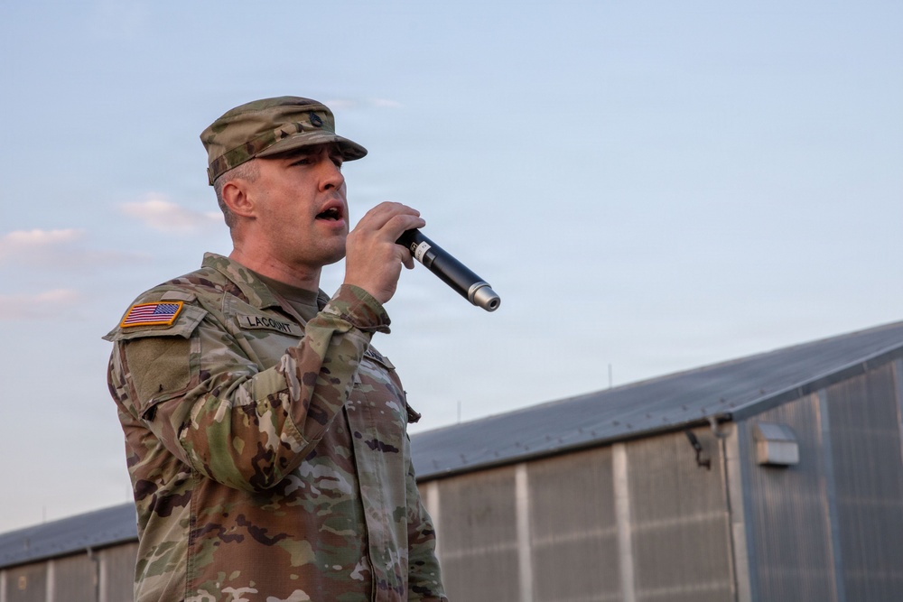A U.S. Army soldier identified by name tape as 'LACOUNT' is speaking or performing into a handheld wireless microphone while wearing OCP (Operational Camouflage Pattern) uniform with a U.S. flag patch on the left shoulder. The setting appears to be an outdoor military installation or base, with large metal structures visible in the background. The image was taken during dusk or early evening based on the soft lighting conditions.