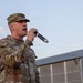 A U.S. Army soldier identified by name tape as 'LACOUNT' is speaking or performing into a handheld wireless microphone while wearing OCP (Operational Camouflage Pattern) uniform with a U.S. flag patch on the left shoulder. The setting appears to be an outdoor military installation or base, with large metal structures visible in the background. The image was taken during dusk or early evening based on the soft lighting conditions.