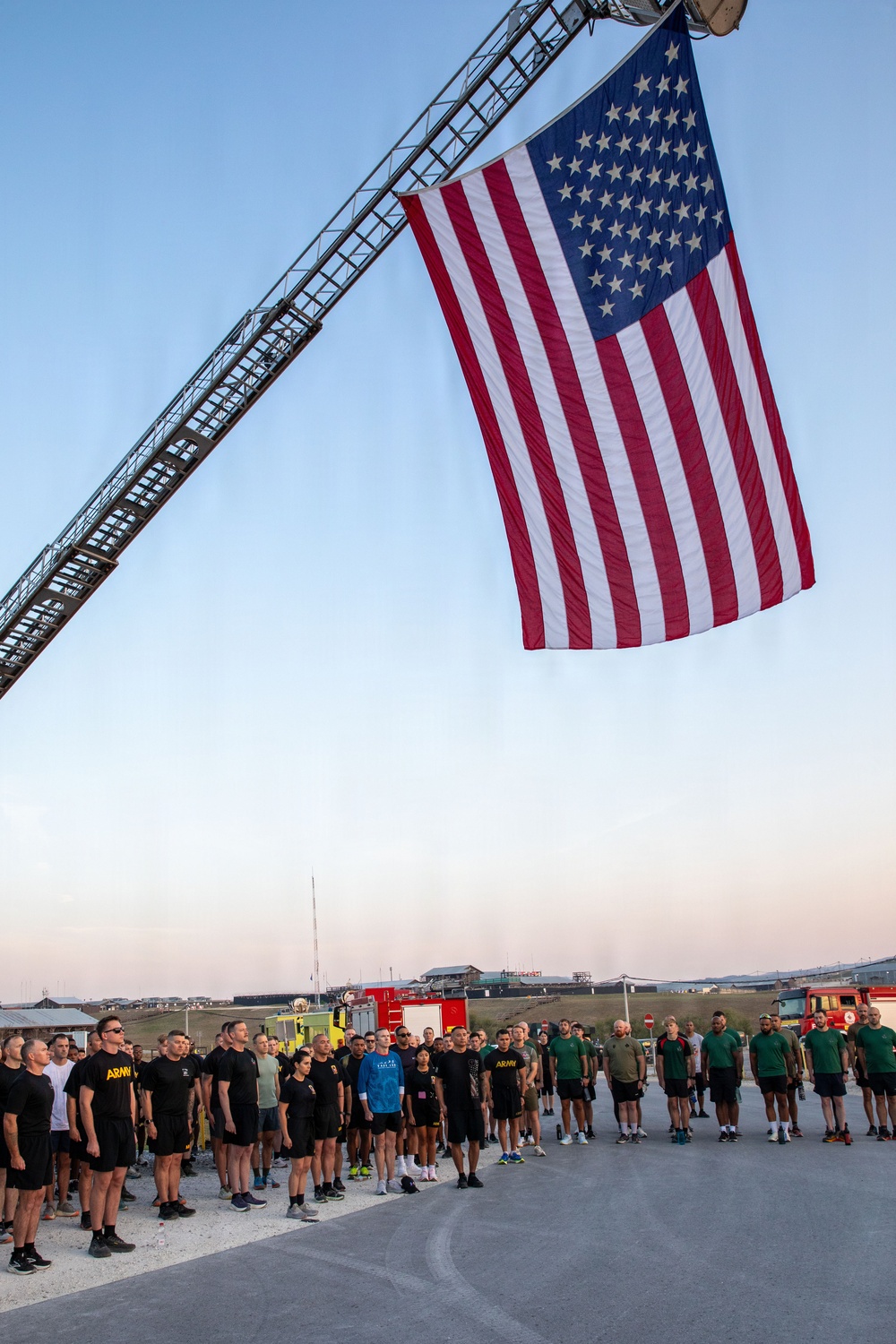 A large American flag is suspended from an extended fire truck ladder over a group of military personnel and civilians standing in formation on a paved surface. The scene appears to take place at dawn or dusk at what looks like a military installation or base, with fire trucks visible in the background. The gathering has the appearance of a memorial or commemorative ceremony, likely a 9/11 tribute event given the combination of military personnel, firefighting equipment, and prominent flag display.