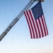 A large American flag is suspended from an extended fire truck ladder over a group of military personnel and civilians standing in formation on a paved surface. The scene appears to take place at dawn or dusk at what looks like a military installation or base, with fire trucks visible in the background. The gathering has the appearance of a memorial or commemorative ceremony, likely a 9/11 tribute event given the combination of military personnel, firefighting equipment, and prominent flag display.
