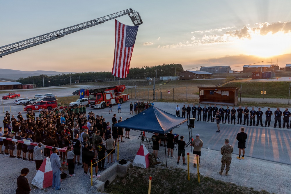 An outdoor memorial or commemorative ceremony is taking place at what appears to be a military installation, with a large American flag suspended vertically from an extended fire truck ladder aerial boom. A large crowd of participants, including military personnel and civilians in dark athletic clothing, are gathered around a blue canopy tent at sunrise or sunset. The presence of a Kosovo flag on a fire truck and the ceremonial formation suggest this may be a 9/11 remembrance event held at a NATO or U.S. military base.