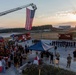 An outdoor memorial or commemorative ceremony is taking place at what appears to be a military installation, with a large American flag suspended vertically from an extended fire truck ladder aerial boom. A large crowd of participants, including military personnel and civilians in dark athletic clothing, are gathered around a blue canopy tent at sunrise or sunset. The presence of a Kosovo flag on a fire truck and the ceremonial formation suggest this may be a 9/11 remembrance event held at a NATO or U.S. military base.
