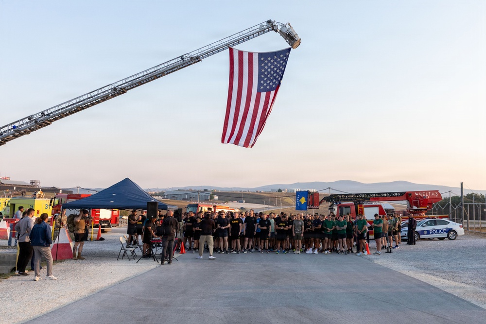 An outdoor ceremony or event is taking place at what appears to be a military or emergency services installation, with a large American flag suspended from a fire truck aerial ladder. A large group of people, many wearing dark military-style PT (physical training) uniforms, are assembled beneath the flag. A Kosovo flag is also visible in the background, suggesting a joint or multinational event.