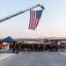 An outdoor ceremony or event is taking place at what appears to be a military or emergency services installation, with a large American flag suspended from a fire truck aerial ladder. A large group of people, many wearing dark military-style PT (physical training) uniforms, are assembled beneath the flag. A Kosovo flag is also visible in the background, suggesting a joint or multinational event.