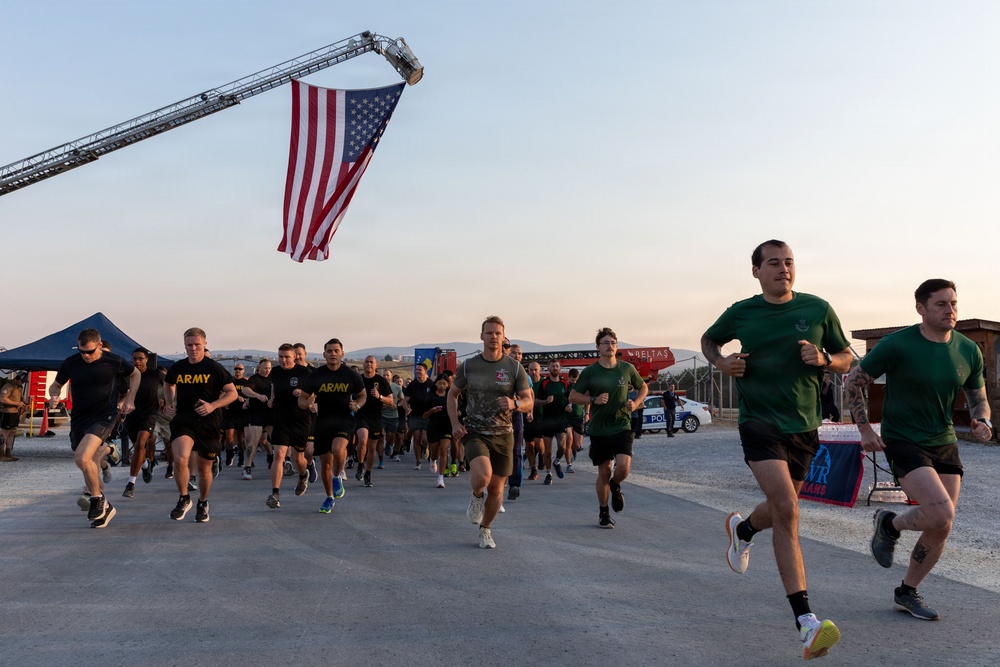 A large group of military personnel and civilians participate in an organized run or physical training event at what appears to be a military installation. A large American flag is suspended from a fire truck ladder boom overhead, suggesting a ceremonial or commemorative event. Participants wear a mix of U.S. Army PT uniforms and civilian athletic wear.