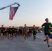 A large group of military personnel and civilians participate in an organized run or physical training event at what appears to be a military installation. A large American flag is suspended from a fire truck ladder boom overhead, suggesting a ceremonial or commemorative event. Participants wear a mix of U.S. Army PT uniforms and civilian athletic wear.