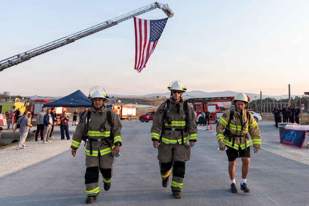 Three firefighters in full turnout gear and white helmets walk toward the camera on a paved surface beneath a large American flag suspended from an aerial ladder truck. The setting appears to be an outdoor firefighter event or memorial exercise, with fire trucks, a blue canopy tent, and a crowd of onlookers visible in the background. The warm, low-angle lighting suggests early morning or late afternoon.