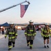 Three firefighters in full turnout gear and white helmets walk toward the camera on a paved surface beneath a large American flag suspended from an aerial ladder truck. The setting appears to be an outdoor firefighter event or memorial exercise, with fire trucks, a blue canopy tent, and a crowd of onlookers visible in the background. The warm, low-angle lighting suggests early morning or late afternoon.