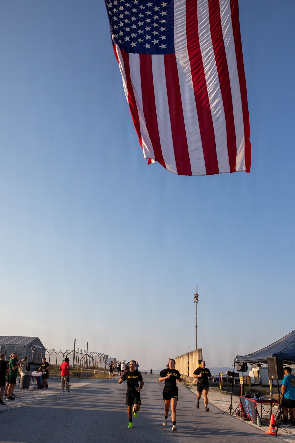 Three U.S. Army soldiers in black PT (physical training) uniforms run along a paved path beneath a large American flag suspended overhead. The setting appears to be a military installation or base, with security fencing, temporary structures, and event tents visible in the background. The scene suggests an organized fitness run or commemorative race event.