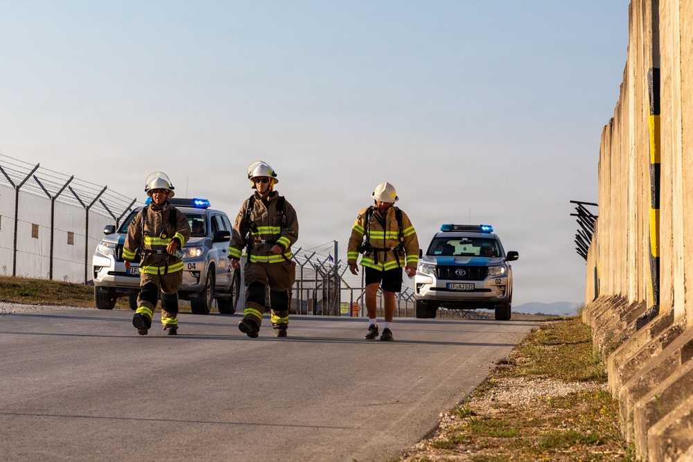 Three individuals wearing firefighter gear and white helmets are walking along a road near a secured perimeter fence. Two Toyota SUVs with blue emergency lights activated follow behind them. The setting appears to be a secured facility, possibly an airport or military installation, based on the barbed wire fencing and concrete barriers.