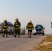 Three individuals wearing firefighter gear and white helmets are walking along a road near a secured perimeter fence. Two Toyota SUVs with blue emergency lights activated follow behind them. The setting appears to be a secured facility, possibly an airport or military installation, based on the barbed wire fencing and concrete barriers.