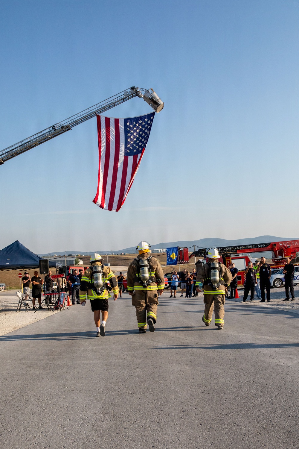 Three firefighters in full gear including SCBA tanks and helmets walk toward a large American flag suspended from a fire truck aerial ladder. The scene appears to be an outdoor ceremonial or memorial event, likely a 9/11 tribute or firefighter competition, with spectators, fire trucks, and a blue tent visible in the background. A Kosovo flag is also visible near the fire trucks, suggesting an international or NATO-related setting.