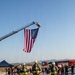 Three firefighters in full gear including SCBA tanks and helmets walk toward a large American flag suspended from a fire truck aerial ladder. The scene appears to be an outdoor ceremonial or memorial event, likely a 9/11 tribute or firefighter competition, with spectators, fire trucks, and a blue tent visible in the background. A Kosovo flag is also visible near the fire trucks, suggesting an international or NATO-related setting.