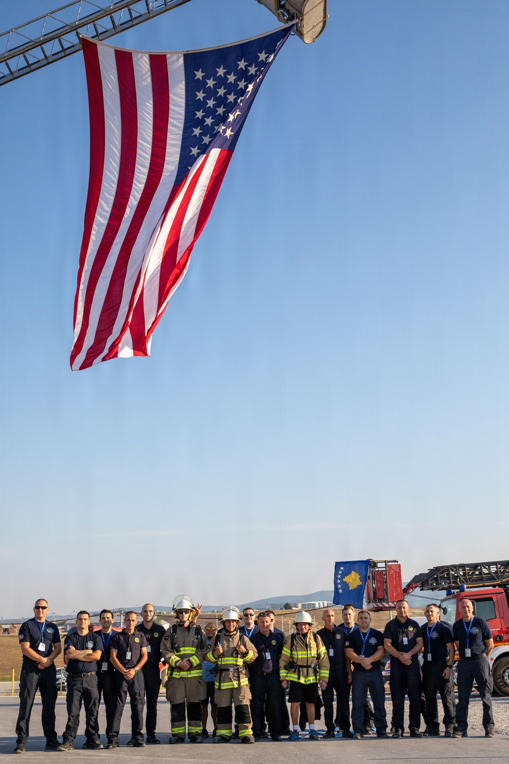A group of approximately 16 firefighters and emergency personnel pose for a group photo beneath a large American flag suspended from a fire truck aerial ladder. Several individuals are wearing full firefighting turnout gear with white helmets, while others are dressed in dark navy uniforms with blue lanyards/badges. A fire truck with what appears to be a Kosovo flag emblem is visible in the background, suggesting an international firefighting cooperation or training event.
