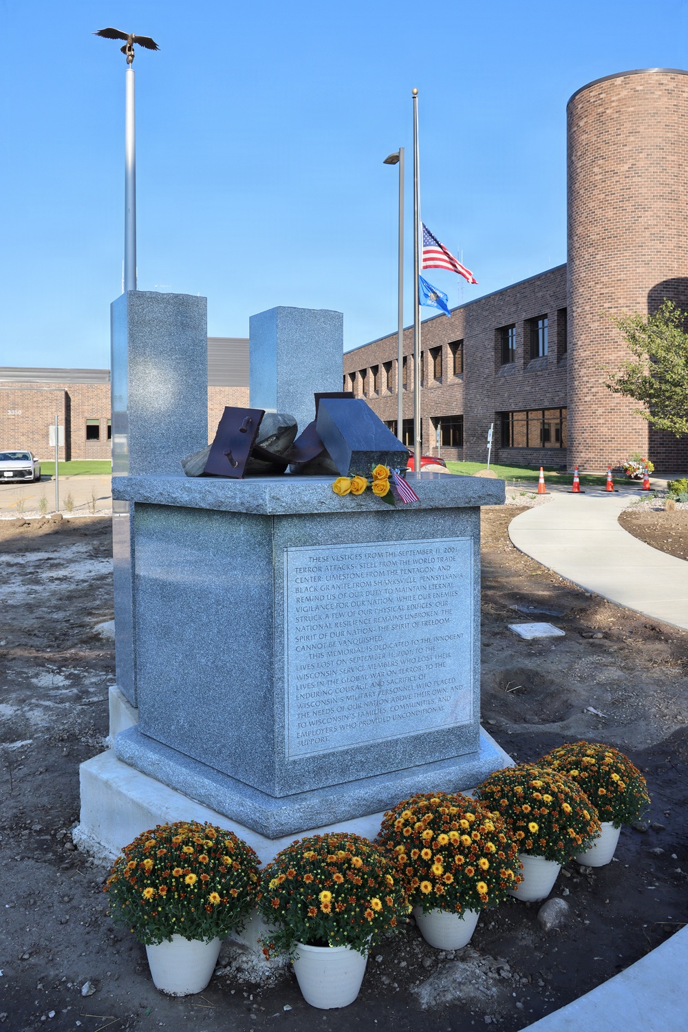 A 9/11 memorial monument made of gray granite is displayed in front of a brick building, featuring twisted steel remnants and stone fragments atop the pedestal. The monument includes an inscribed dedication panel referencing the September 11, 2001 terror attacks and mentions Wisconsin service members. The setting includes potted chrysanthemums at the base, yellow roses placed on top, and flags flying at half-staff in the background.