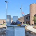 A 9/11 memorial monument made of gray granite is displayed in front of a brick building, featuring twisted steel remnants and stone fragments atop the pedestal. The monument includes an inscribed dedication panel referencing the September 11, 2001 terror attacks and mentions Wisconsin service members. The setting includes potted chrysanthemums at the base, yellow roses placed on top, and flags flying at half-staff in the background.