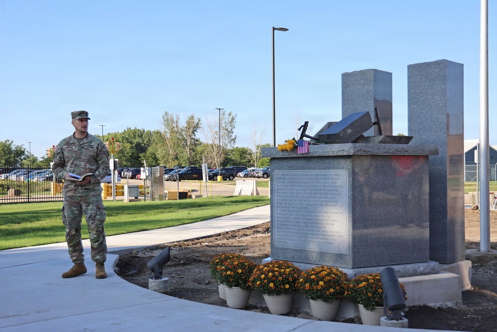 A U.S. Army soldier in OCP (Operational Camouflage Pattern) uniform stands beside a granite 9/11 memorial monument, appearing to read from a book during a commemorative ceremony. The memorial features two tall granite pillars representing the Twin Towers, with what appears to be a steel beam relic and a small American flag placed on top. The setting appears to be a military installation with a parking lot and trees visible in the background.