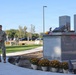 A U.S. Army soldier in OCP (Operational Camouflage Pattern) uniform stands beside a granite 9/11 memorial monument, appearing to read from a book during a commemorative ceremony. The memorial features two tall granite pillars representing the Twin Towers, with what appears to be a steel beam relic and a small American flag placed on top. The setting appears to be a military installation with a parking lot and trees visible in the background.