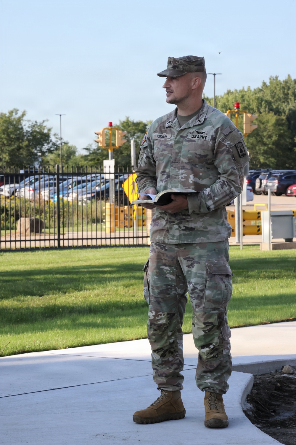 A U.S. Army soldier named Hanson, wearing Operational Camouflage Pattern (OCP) uniform, stands on a concrete walkway holding an open book, appearing to be speaking or reading aloud. He wears the rank insignia of a Lieutenant Colonel (oak leaf) on his collar and a chaplain cross insignia, suggesting he may be a military chaplain. The setting appears to be a military installation entrance or gate area during daytime.