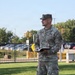A U.S. Army soldier named Hanson, wearing Operational Camouflage Pattern (OCP) uniform, stands on a concrete walkway holding an open book, appearing to be speaking or reading aloud. He wears the rank insignia of a Lieutenant Colonel (oak leaf) on his collar and a chaplain cross insignia, suggesting he may be a military chaplain. The setting appears to be a military installation entrance or gate area during daytime.