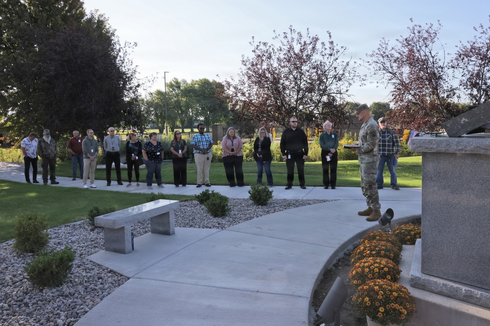 An outdoor memorial or commemorative ceremony is taking place in a well-maintained garden setting. A person in military uniform appears to be speaking or presiding at a stone monument or memorial, while approximately 15-20 civilians stand attentively in a semicircle listening. The setting includes decorative landscaping with stone benches, chrysanthemums, gravel beds, and mature trees suggesting an autumn season.