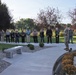 An outdoor memorial or commemorative ceremony is taking place in a well-maintained garden setting. A person in military uniform appears to be speaking or presiding at a stone monument or memorial, while approximately 15-20 civilians stand attentively in a semicircle listening. The setting includes decorative landscaping with stone benches, chrysanthemums, gravel beds, and mature trees suggesting an autumn season.