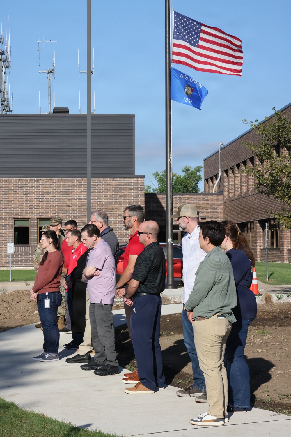 A group of approximately 12-15 civilians and at least one military personnel are standing at attention facing a flagpole displaying the United States flag and the Wisconsin state flag (dated 1848). The gathering appears to be a formal flag ceremony or dedication event at what looks like a Wisconsin National Guard or government facility with brick buildings in the background. The setting suggests an official outdoor ceremony, possibly a building dedication, flag raising, or patriotic observance.