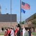 A group of approximately 12-15 civilians and at least one military personnel are standing at attention facing a flagpole displaying the United States flag and the Wisconsin state flag (dated 1848). The gathering appears to be a formal flag ceremony or dedication event at what looks like a Wisconsin National Guard or government facility with brick buildings in the background. The setting suggests an official outdoor ceremony, possibly a building dedication, flag raising, or patriotic observance.