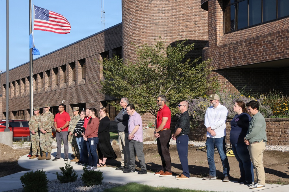 A group of approximately 15-20 people, including several individuals in military camouflage uniforms and civilians, are gathered outdoors in front of a large brick building. They appear to be participating in a formal outdoor ceremony near a flagpole flying a full-sized American flag and a smaller blue flag. The setting suggests a government, military, or institutional facility.