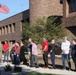 A group of approximately 15-20 people, including several individuals in military camouflage uniforms and civilians, are gathered outdoors in front of a large brick building. They appear to be participating in a formal outdoor ceremony near a flagpole flying a full-sized American flag and a smaller blue flag. The setting suggests a government, military, or institutional facility.