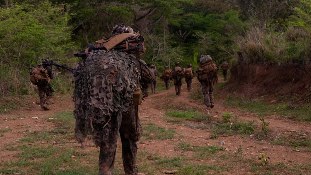 A group of military personnel in full combat gear are patrolling along a dirt trail through a tropical or subtropical environment. The foreground soldier is wearing a ghillie suit or camouflage netting over their pack, suggesting a sniper or reconnaissance role. The unit appears to be conducting a field training exercise or tactical movement through rugged terrain.