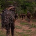 A group of military personnel in full combat gear are patrolling along a dirt trail through a tropical or subtropical environment. The foreground soldier is wearing a ghillie suit or camouflage netting over their pack, suggesting a sniper or reconnaissance role. The unit appears to be conducting a field training exercise or tactical movement through rugged terrain.