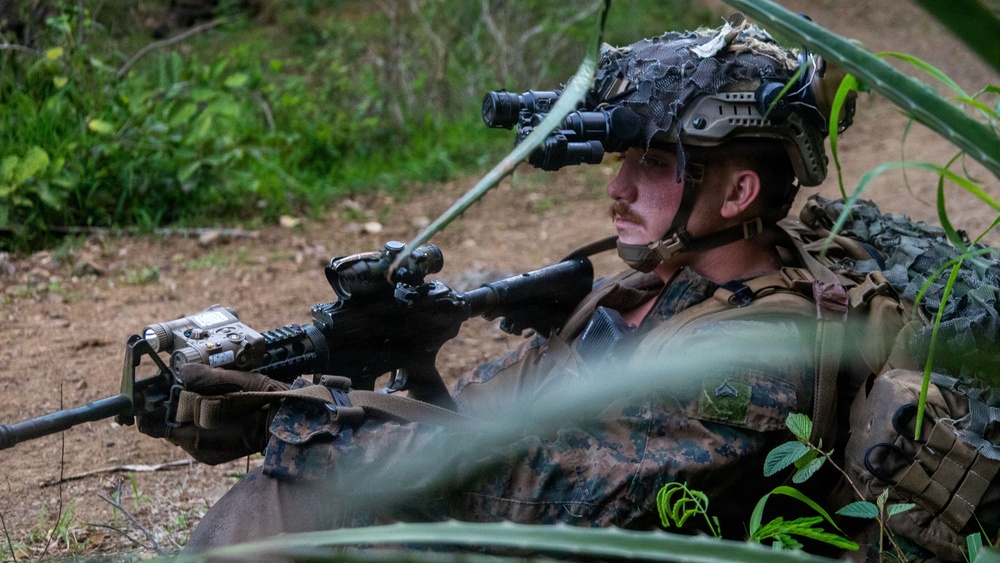 A U.S. Marine in full combat gear is positioned in a prone or low-crouch stance in a wooded outdoor environment, holding an assault rifle with optical attachments. The Marine is wearing a tactical helmet equipped with night vision goggles (NVGs) flipped down over one eye, along with camouflage netting and full battle kit. The image appears to be taken during a training exercise based on the controlled setting and visible rank insignia.