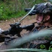 A U.S. Marine in full combat gear is positioned in a prone or low-crouch stance in a wooded outdoor environment, holding an assault rifle with optical attachments. The Marine is wearing a tactical helmet equipped with night vision goggles (NVGs) flipped down over one eye, along with camouflage netting and full battle kit. The image appears to be taken during a training exercise based on the controlled setting and visible rank insignia.