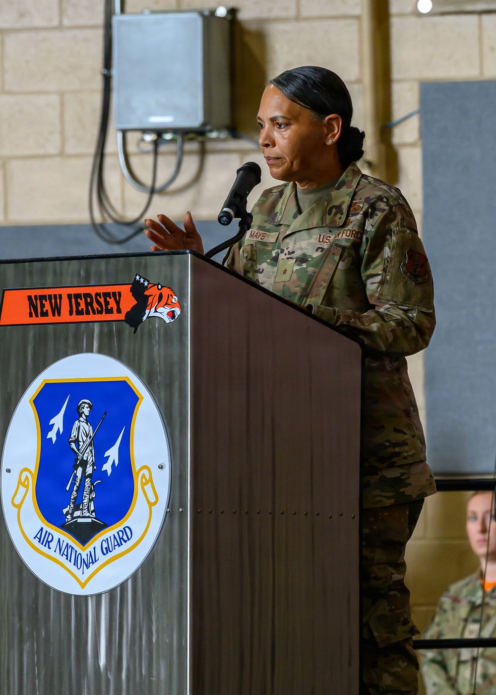 A U.S. Air Force officer with the name tape 'MAYS' and a single star insignia (Brigadier General) is speaking at a podium during an official ceremony. The podium displays the New Jersey Air National Guard emblem along with a tiger mascot graphic and the state outline. A second uniformed service member is partially visible in the background.