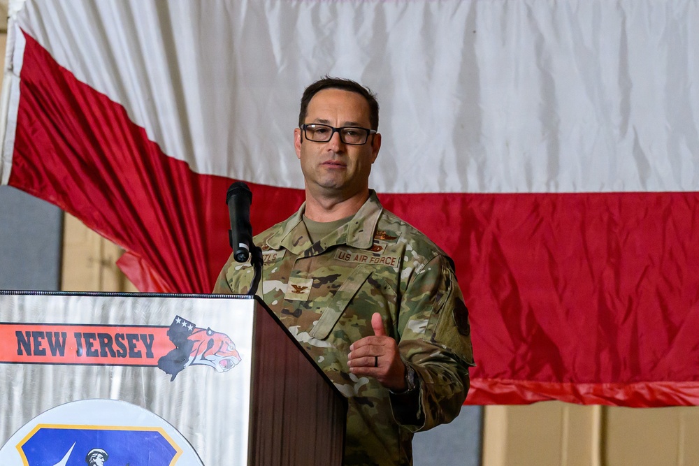 A U.S. Air Force officer in OCP (Operational Camouflage Pattern) uniform is speaking at a podium during an official military ceremony or event. The podium displays a 'New Jersey' insignia with what appears to be a tiger emblem, suggesting this is a New Jersey Air National Guard event. A large red and white flag, consistent with the Polish national flag, is displayed in the background.