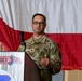 A U.S. Air Force officer in OCP (Operational Camouflage Pattern) uniform is speaking at a podium during an official military ceremony or event. The podium displays a 'New Jersey' insignia with what appears to be a tiger emblem, suggesting this is a New Jersey Air National Guard event. A large red and white flag, consistent with the Polish national flag, is displayed in the background.