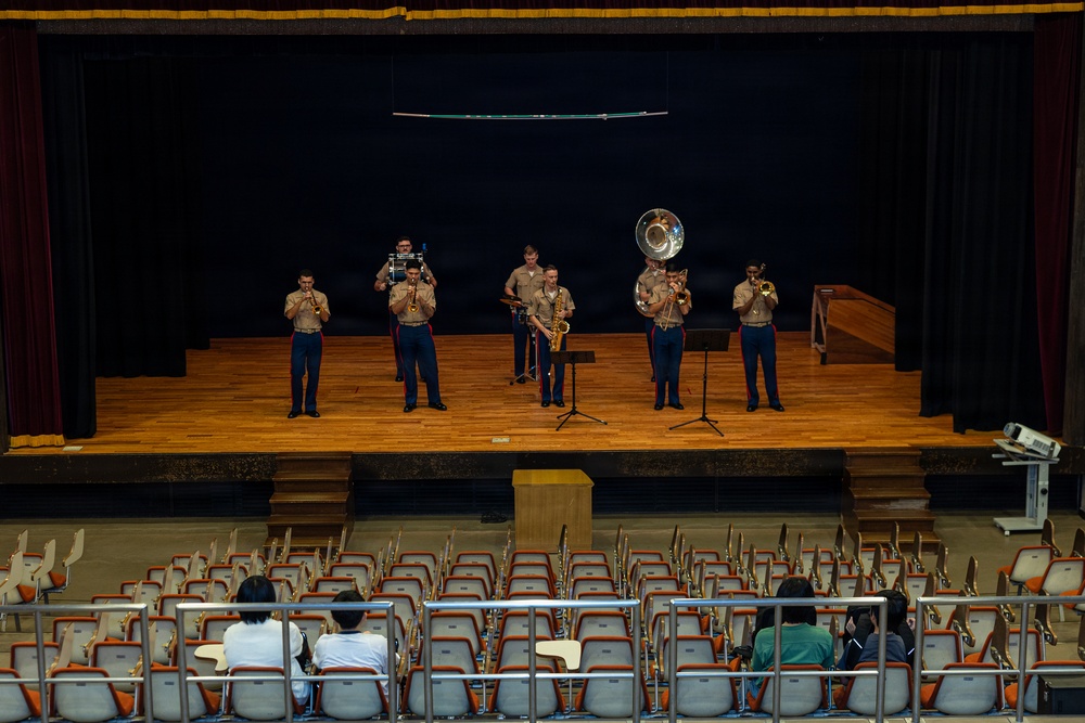 A small military band ensemble, appearing to be U.S. Marines in tan and blue dress uniforms, performs on a wooden auditorium stage. The group consists of approximately six musicians playing brass and woodwind instruments to a sparse audience of roughly four seated spectators. The setting appears to be a school or community auditorium with rows of mostly empty orange seats.
