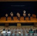 A small military band ensemble, appearing to be U.S. Marines in tan and blue dress uniforms, performs on a wooden auditorium stage. The group consists of approximately six musicians playing brass and woodwind instruments to a sparse audience of roughly four seated spectators. The setting appears to be a school or community auditorium with rows of mostly empty orange seats.